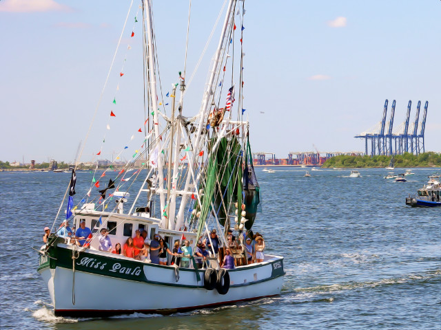 A shrimp boat moving on the Cooper River at the Blessing of the Fleet boat parade. There are many people on the boat, which is painted white with green trim. Along the side of the boat is its name, Miss Paula.