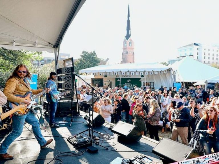View from the stage of a rock band playing live with an audience and tents in the background, and a view of the Charleston skyline with a large church steeple