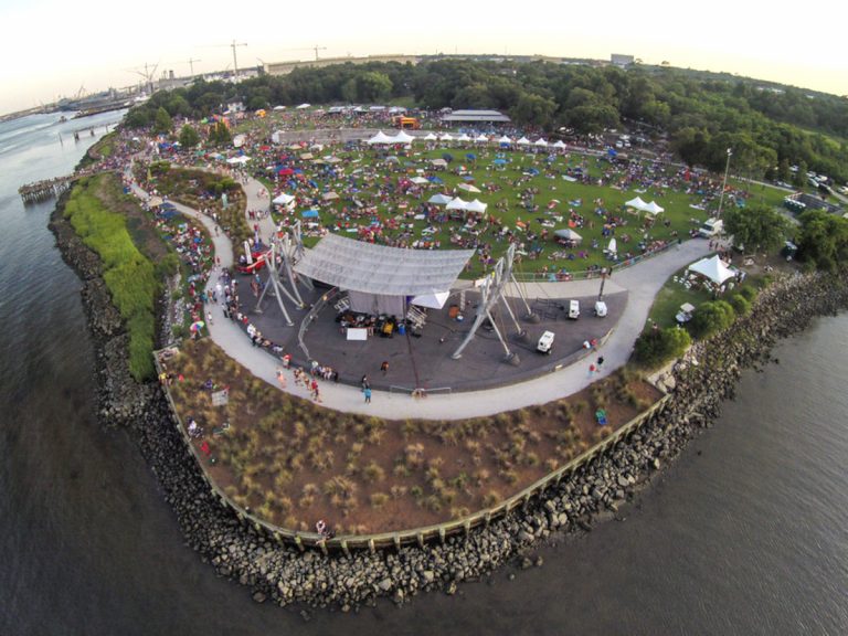 An aerial fisheye lens view of Riverfront Park in Charleston, with the park area and concert stage in the center, surrounded by coastal water views of the surrounding river along the bottom