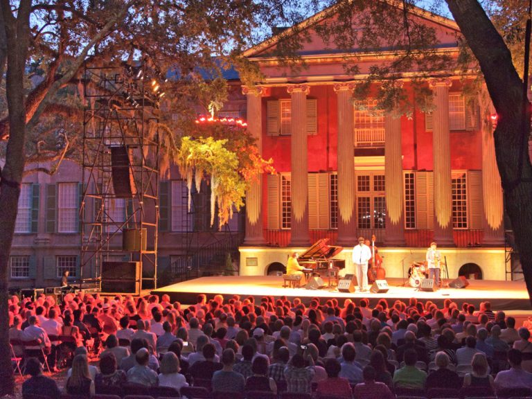 An outdoor concert at the College of Charleston Randolph Hall during the Spoleto Festival. The band is performing on a stage with the tall columns of Randolph Hall behind them and a crowd of onlookers bathed in an red-orange tinted light.