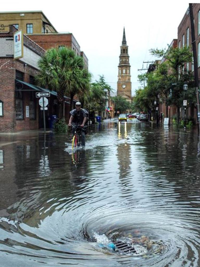 A flooded street in downtown Charleston with stormwater flowing into a drain, with the steeple of St. Philips Church in the background