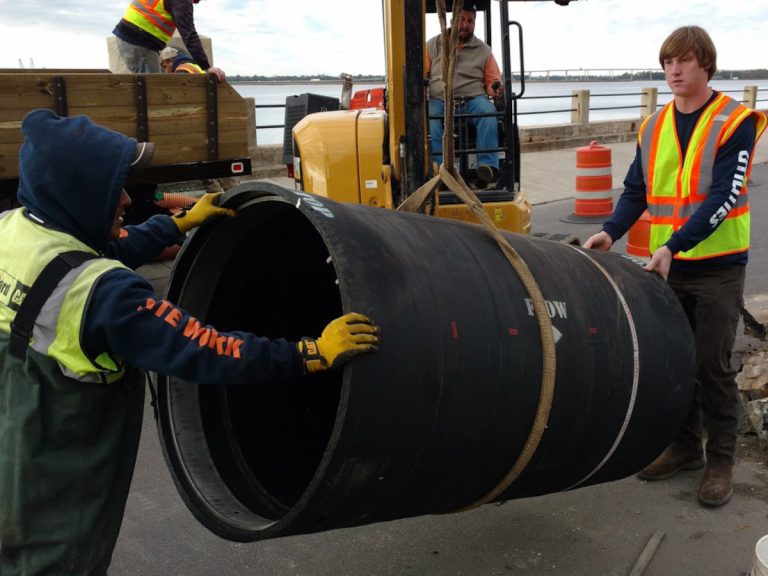 Workers handling a large black pipe containing a check valve, preparing to install it under a street in Charleston
