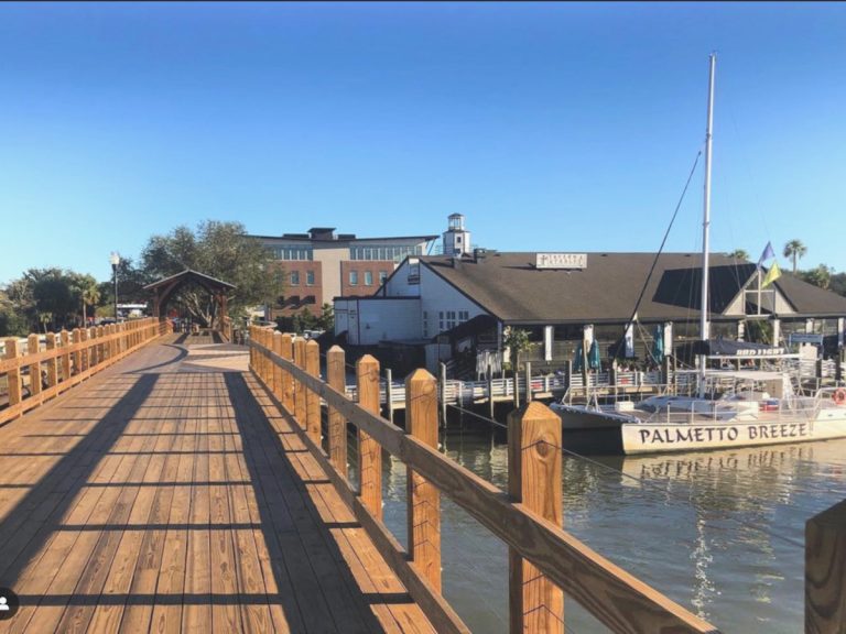 The wooden boardwalk of the Shem Creek Bridge in Mount Pleasant with a view of a restaurant on the right with a catamaran boat docked.