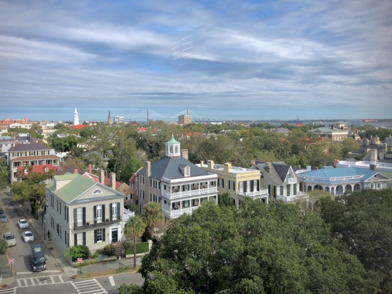 The skyline of Charleston as viewed from South Battery Street, showing grand homes facing large green oak trees against a blue sky with clouds.
