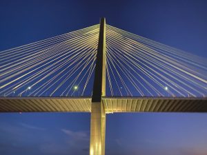 An upward view of one tower of the Ravenel Bridge with lights shining on the tower and cables against a dark blue sky.