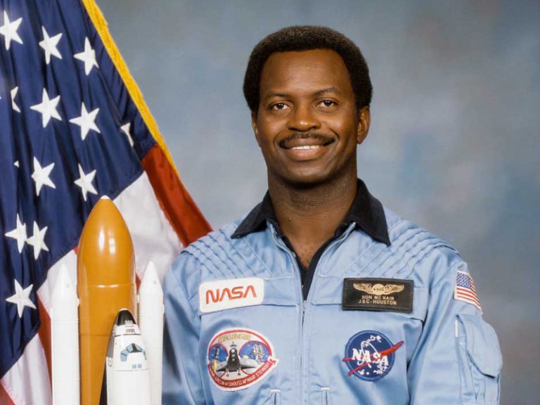 A photo of astronaut Dr. Ron McNair standing next to a model of the Space Shuttle with an American flag behind him