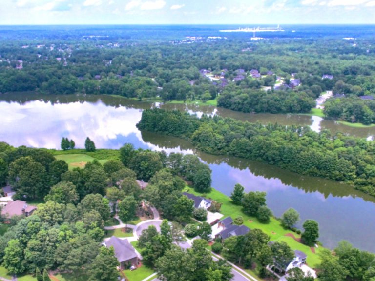 An aerial view of the Crowfield neighborhood showing homes with green lawns and trees along a waterway reflecting the blue sky and clouds.